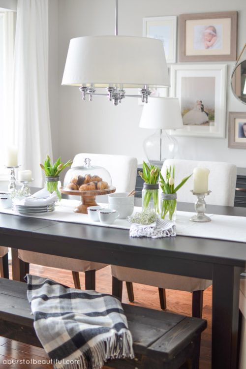 An open-concept dining room and living room is bright and inviting with a large farmhouse table, grey couch and eclectic gallery wall | A Burst of Beautiful