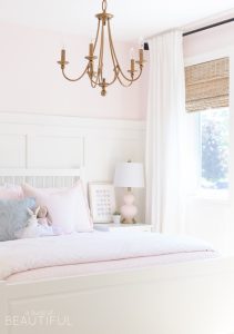 This sweet pink and white little girl's bedroom is full of soft colors and whimsical details, like board and batten, woven wood shades, a gold chandelier and pale pink bedding.