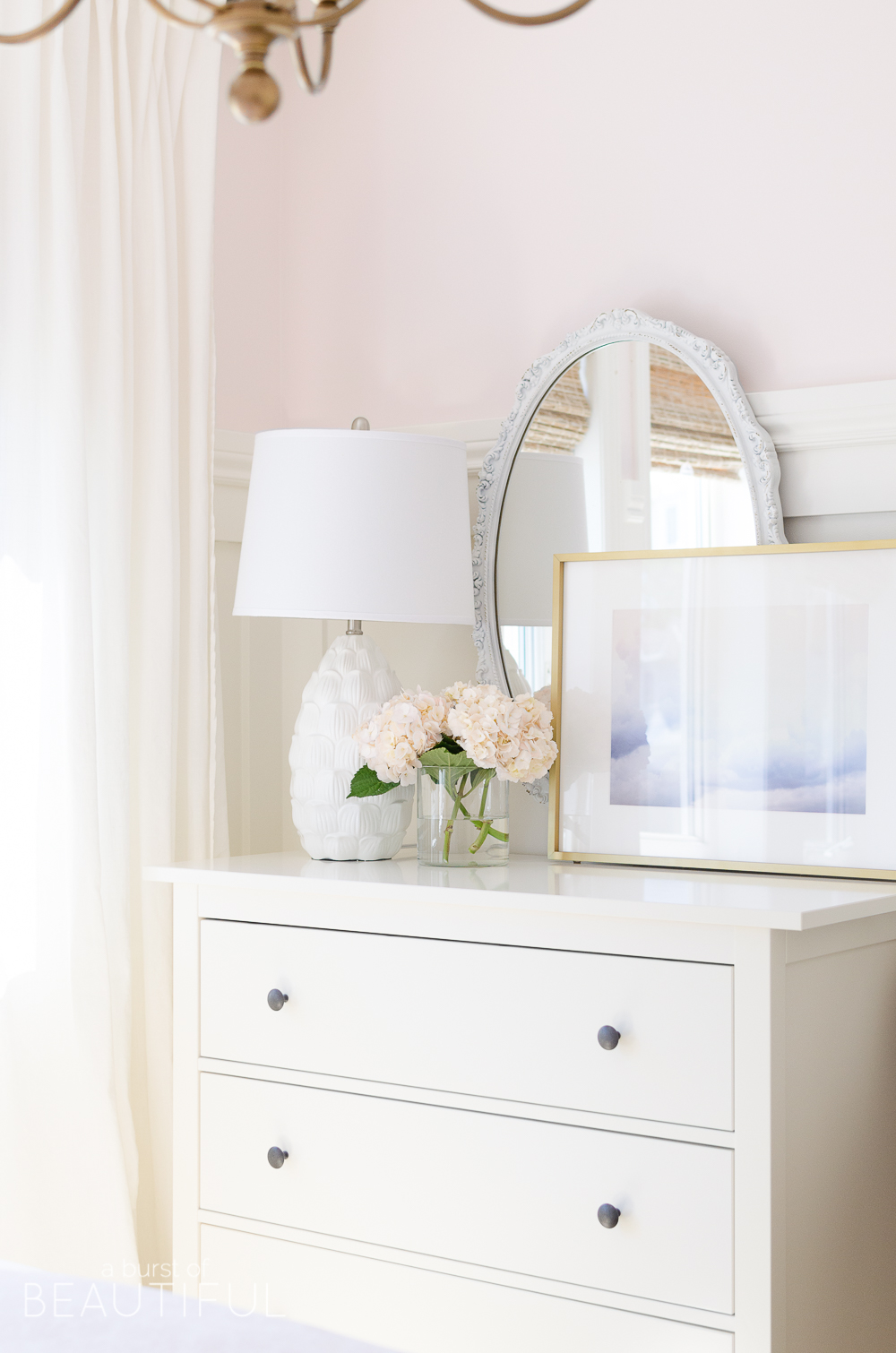 Este doce quarto de menina rosa e branco está cheio de cores suaves e detalhes caprichosos, como tábua e ripa, tons de madeira tecidos, um lustre dourado e roupa de cama rosa pálido.'s bedroom is full of soft colors and whimsical details, like board and batten, woven wood shades, a gold chandelier and pale pink bedding.