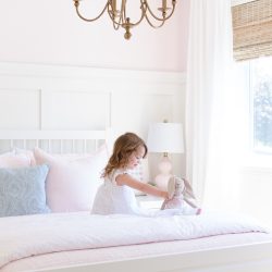This sweet pink and white little girl's bedroom is full of soft colors and whimsical details, like board and batten, woven wood shades, a gold chandelier and pale pink bedding.