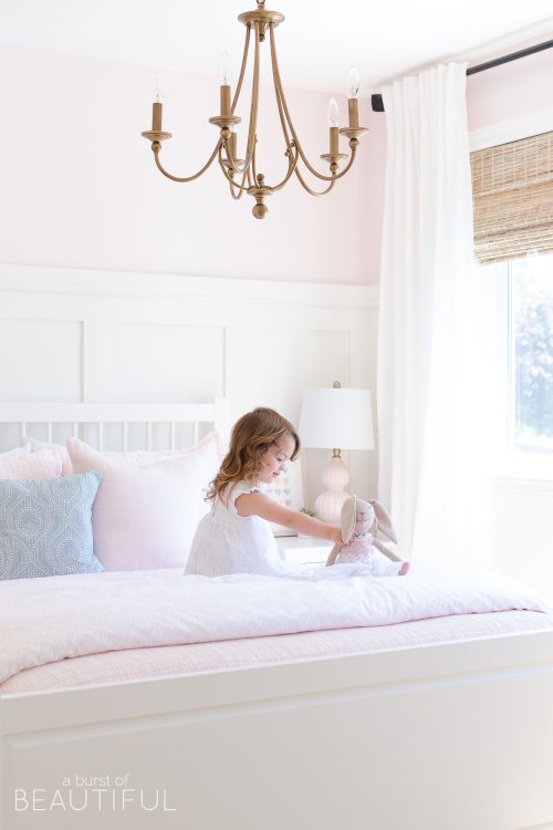This sweet pink and white little girl's bedroom is full of soft colors and whimsical details, like board and batten, woven wood shades, a gold chandelier and pale pink bedding.